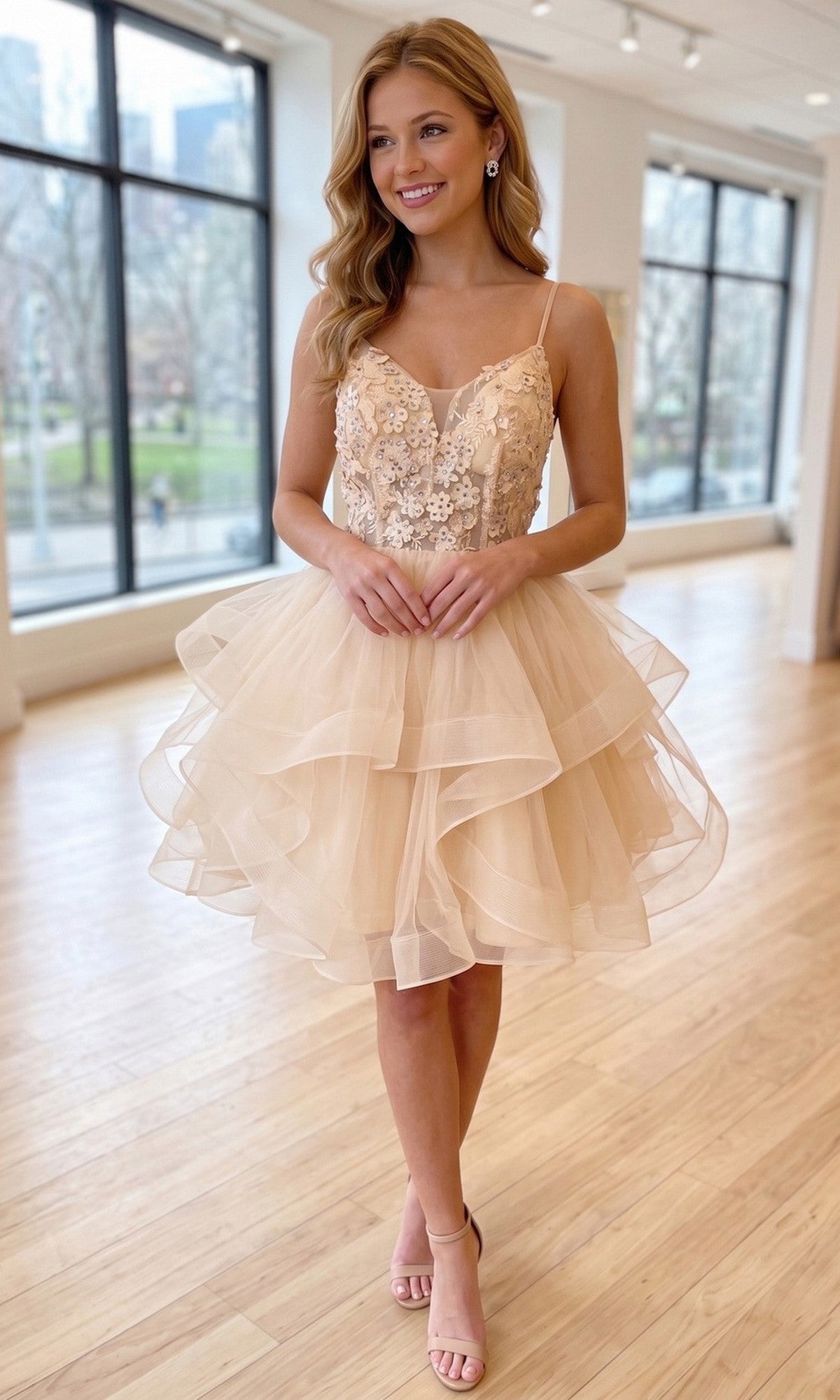 A woman stands indoors on a light wood floor, smiling with a city view visible through large windows behind her, wearing the Chicas C320 Short Homecoming Dress in champagne gold—a sleeveless, layered tulle fit-and-flare design featuring an embroidered floral appliqué bodice.